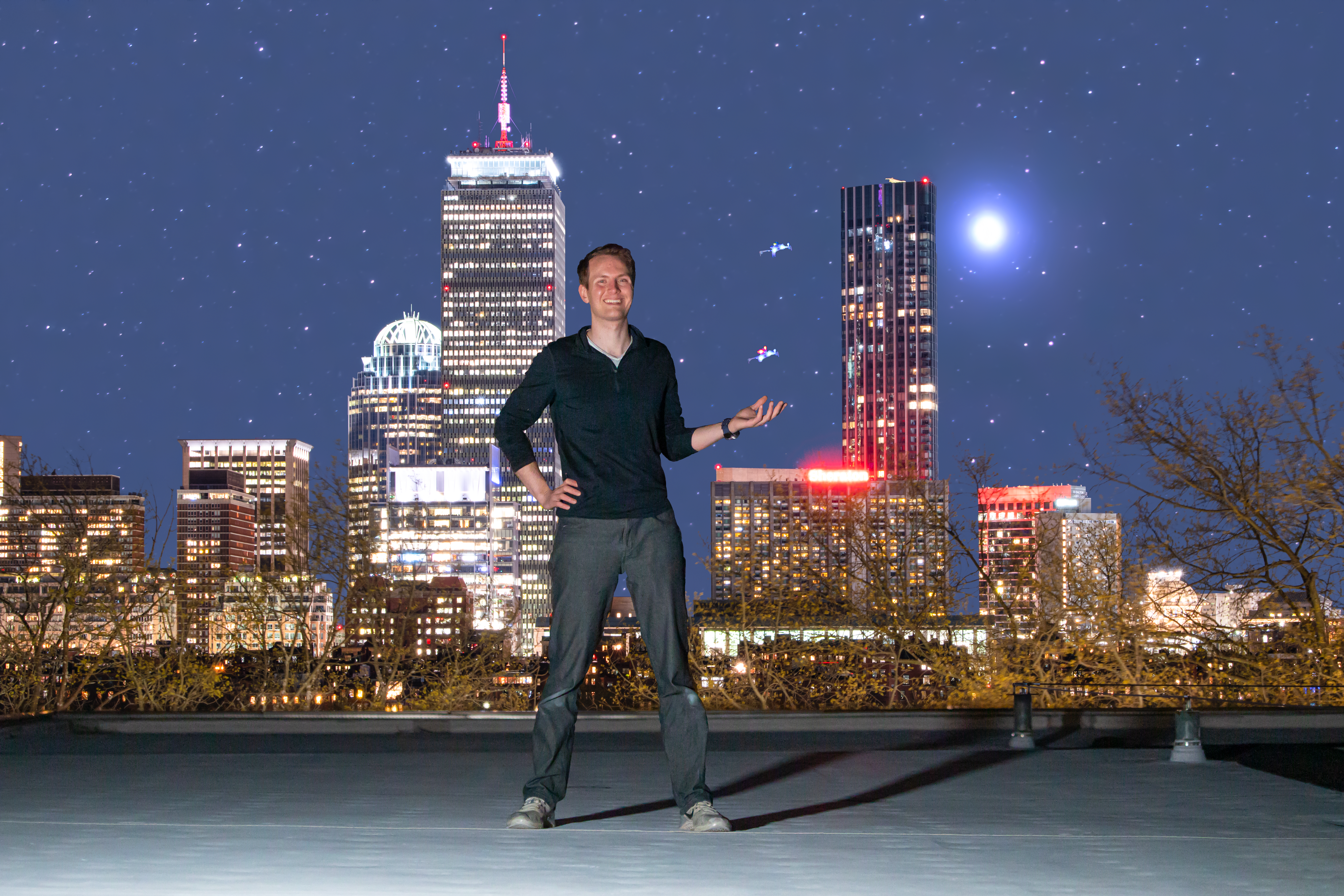 Charles Dawson stands on a roof in the center of the frame. His left hand is extended, and two palm-sized drones fly above it. In the background, stars can be seen in the night sky, and the Boston skyline with the Prudential Center is lit up.