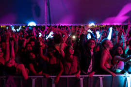 In a darkened tent with red and purple light, hundreds of MIT community members dance with their hands in the air.