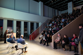 A large crowd facing the well lit panelists in the MIT Museum's stadium seating event venue