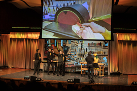 Members of the Gold Team wears black suits and yellow ties on stage, with a screen behind them showing their disc sander in action and a tool wall.