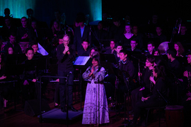 Djuena Tikuna stands on stage in front of many MIT musicians, with her hands pressed together in front of her heart.