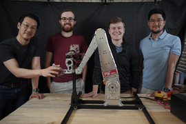  Sangbae Kim, Elijah Stanger-Jones, Andrew SaLoutos, and Hongmin Kim pose for a portrait as they stand behind a metal robot gripper that resembles an excavator. Sangbae Kim’s fingers lace with the gripper.