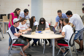 Six students sit at round table studying together with laptops, while four people look on behind them.