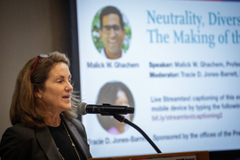 Cynthia Barnhart speaks into a microphone while a screen in background shows Malick Chachem and Tracie D. Jones-Barrett.