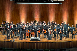 About 50 members of the Orchestra smile for a group photo while on stage and holding their instruments.