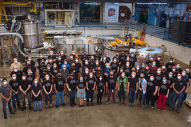 Group of about 70 people wearing face masks stand in front of the cryostat container
