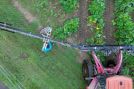 Aerial photo shows two people installing a device on the long arm of a spray boom, while in a field.