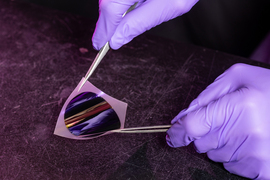 Purple gloved hands and tweezers manipulate a sheet of shiny silicon-type wafer.